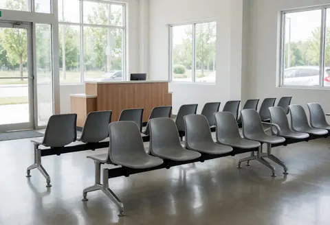 Vue d'un espace d'attente vide et lumineux doté de chaises grises, d'un comptoir en bois et de grandes fenêtres, évoquant un environnement professionnel et épuré.