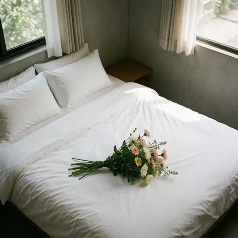 Lit d'hôtel impeccable avec draps blancs, fleurs fraîches dans un vase, lumière naturelle par la fenêtre et murs en béton.
