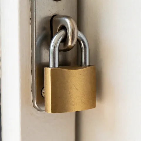 Détail d'un cadenas robuste en métal bronze fixé à un cadre métallique sur un mur blanc, évoquant la protection et la sécurité des biens.