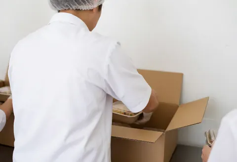 Un travailleur d'une boutique en ligne emballe soigneusement des denrées alimentaires dans une boîte en carton, portant gants et masque dans un environnement propre et bien éclairé.