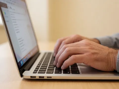 Une main adulte tape sur le clavier d'un ordinateur portable posé sur un bureau en bois, avec un mur beige en arrière-plan et une lumière chaleureuse.