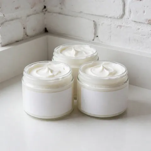 Trois pots de crème blanche avec texture visible sur un plan de travail blanc, mur de briques blanches, éclairage naturel, produit spa et massage.