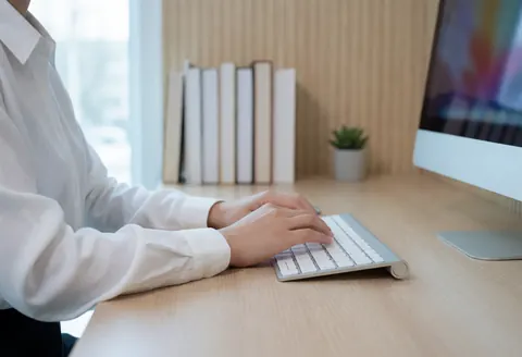 Professionnel tapant au clavier sur un bureau en bois avec livres et plante, dans un environnement de bureau moderne et concentré.