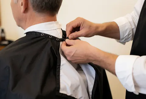 Un barbier ajuste soigneusement la cape protectrice autour du cou d'une cliente dans un salon de coiffure masculin.