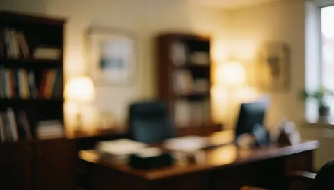 Intérieur d'un bureau professionnel éclairé par une lumière chaude, montrant un bureau en bois, des étagères avec des livres et une plante près d'une fenêtre.
