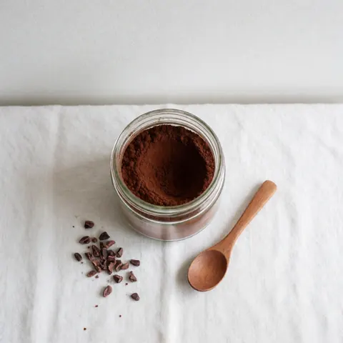 Une jarre en verre remplie de cacao en poudre artisanale accompagnée d'une cuillère en bois, le tout disposé sur un fond blanc avec une lumière douce.