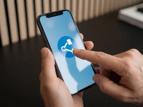 Gros plan sur une main tenant un téléphone portable avec des icônes de partage visibles sur l'écran, posé sur un bureau en bois dans un contexte d'apprentissage en ligne.