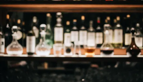 Arrière-plan de bar élégant et sombre mettant en valeur des bouteilles de spiritueux et des décanteurs sur un comptoir en bois avec des lumières floues.