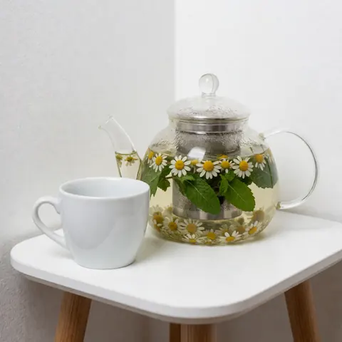 Une théière en verre remplie de camomille et de menthe posée sur une table blanche, accompagnée d'une tasse blanche. Le fond blanc crée une ambiance calme et naturelle.