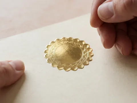 Mains posant un sceau doré sur un certificat en papier crème texturé, avec des détails visibles et un fond beige doux.