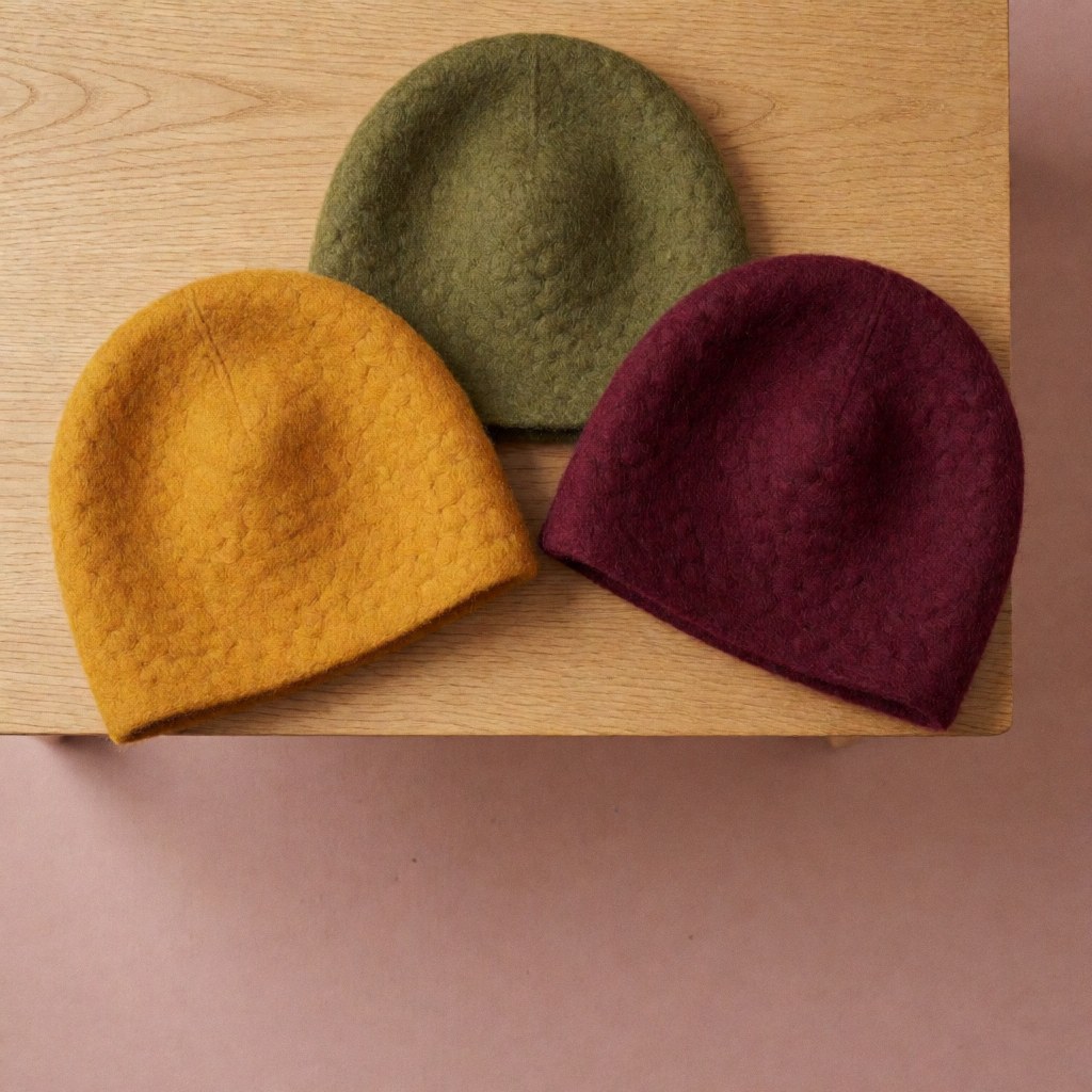 Trois bonnets en laine teints dans des tons chauds disposés en triangle sur une table en bois clair avec un fond rose poudré.