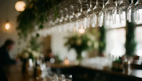 Fond de bar élégant avec des verres à vin suspendus, du feuillage vert et une lumière douce créant une ambiance chaleureuse pour événements.