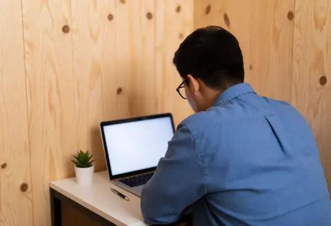 Vue d'une personne concentrée utilisant un ordinateur portable sur un bureau en bois chez elle, avec des plantes succulentes et une lumière douce dans l'obscurité.