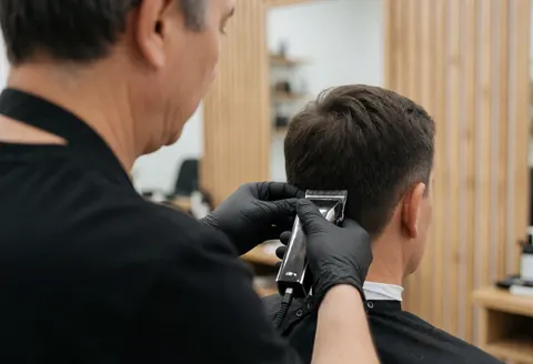 Un barbier portant des gants noirs coupe les cheveux d'un client avec une tondeuse électrique devant un mur en bois.