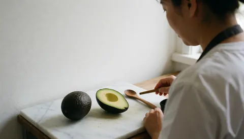 Une cuisinière de dos prépare un avocat frais sur une table en marbre blanc avec une cuillère en bois et une lumière naturelle douce.