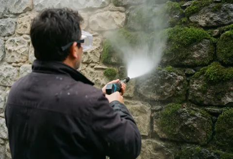 Un ouvrier équipé de lunettes de protection utilise une pistolet à eau pour nettoyer un mur en pierre couvert de mousse, révélant le contraste entre les surfaces sales et propres.