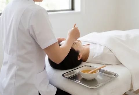 Une thérapeute applique un masque de miel sur le visage d'une cliente dans une clinique esthétique. Ambiance propre, professionnelle et relaxante avec lit, plateau et cuillère.