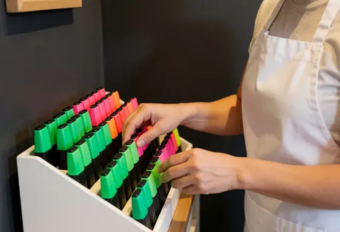 Employé de librairie rangeant des marqueurs colorés sur un présentoir blanc devant un mur gris, avec une lumière chaude et des mains au premier plan.