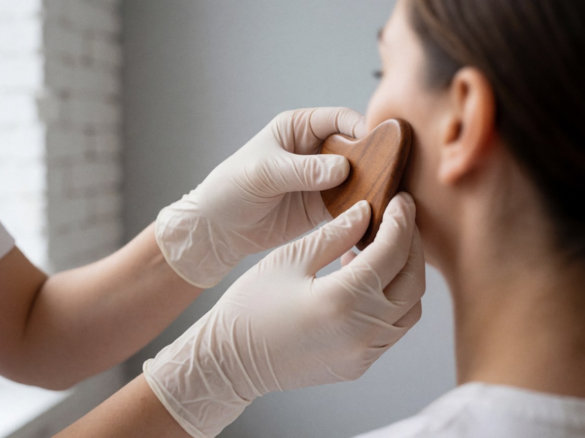 Des mains portant des gants blancs appliquent une technique de massage facial avec un outil en bois sur la joue, devant un mur de briques et un fond gris.