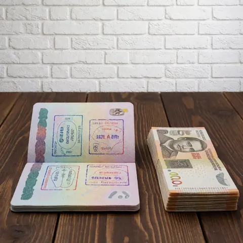 Passeport ouvert révélant des tampons de voyage posé à côté d'une pile de billets de monnaie locale sur une surface en bois sombre.