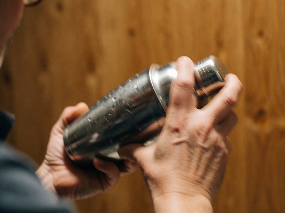 Mains d'un barman secouant vigoureusement un shaker en métal recouvert de gouttelettes de condensation sur un fond en bois chaleureux.