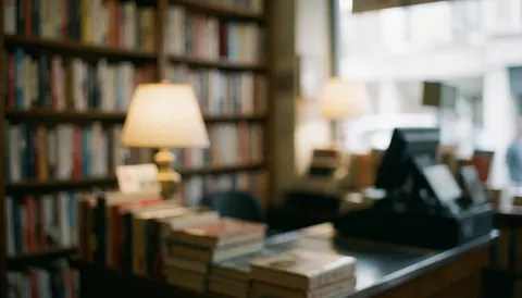 Vue floue d'une caisse en bois dans une librairie avec des étagères de livres et une petite lampe créant une atmosphère calme et chaleureuse.