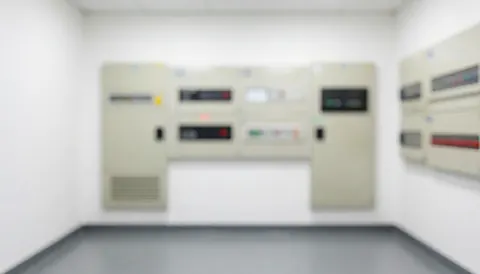Vue de face de panneaux électriques montés sur un mur blanc dans une salle technique avec un sol gris et un éclairage uniforme pour fond professionnel.