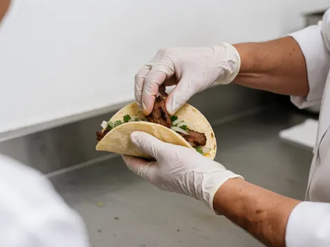 Mains gantées assemblent un taco sur une surface en acier inoxydable, montrant la viande grillée, la tortilla, le cilantro et l'oignon dans un contexte de cuisine professionnelle.