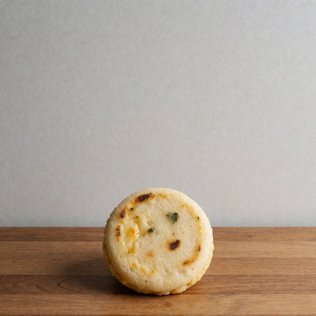 Arepita dorée avec fromage fondu sur une surface en bois, fond gris clair, vue de dessus, style professionnel.