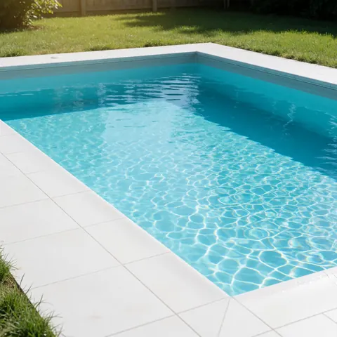 Piscine enterrée récemment terminée avec eau turquoise brillante, carrelage blanc et pelouse verte sous un soleil intense, idéale pour promouvoir des services de construction.