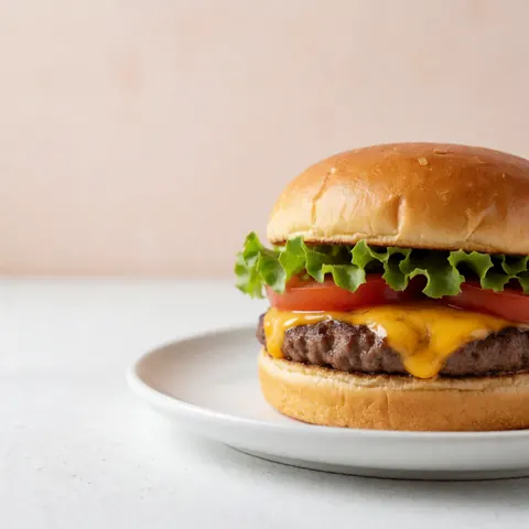 Un hamburger classique avec du fromage fondu, de la laitue et une tranche de tomate, posé sur une assiette blanche devant un mur en terre cuite claire éclairé par la lumière naturelle.