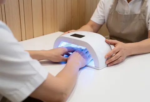 Une manucure professionnelle utilise une lampe blanche pour durcir le gel sur les ongles d'un client dans un salon moderne aux murs en bois clair.