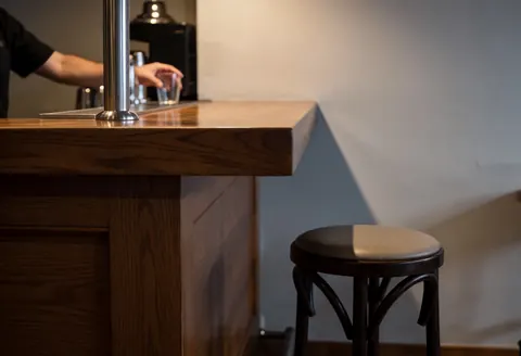 Barre en bois poli avec une chaise de bar au premier plan, mur gris en arrière-plan, main de barman en arrière-plan, éclairage chaleureux et atmosphérique.