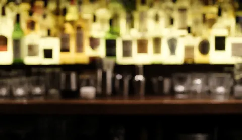 Arrière-plan de bar avec des bouteilles et des verres flous, éclairage chaleureux, idéal pour texte publicitaire ou web.