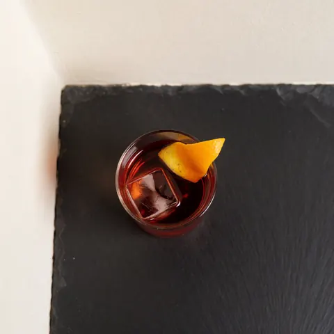 Vue aérienne d'un verre contenant un cocktail rouge foncé avec des glaçons et une tranche d'orange sur une surface sombre.