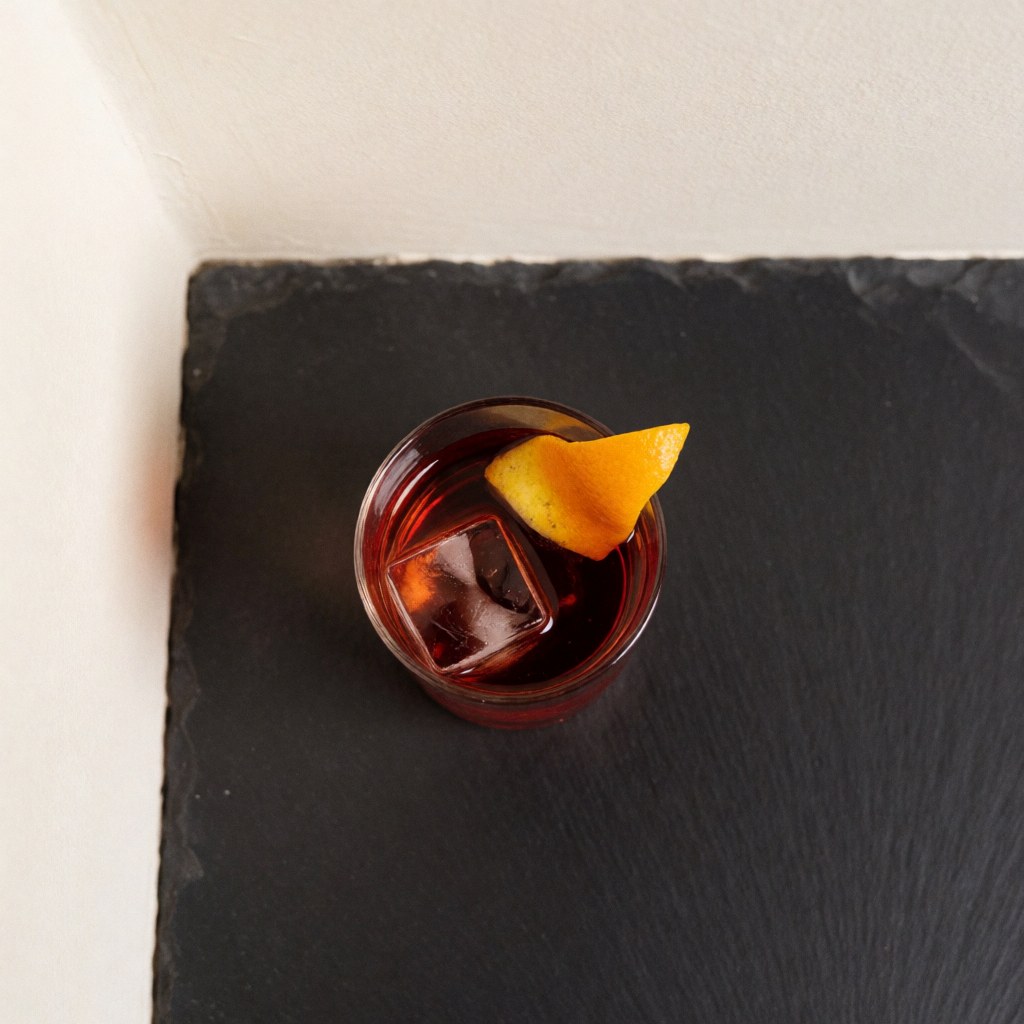 Vue aérienne d'un verre contenant un cocktail rouge foncé avec des glaçons et une tranche d'orange sur une surface sombre.