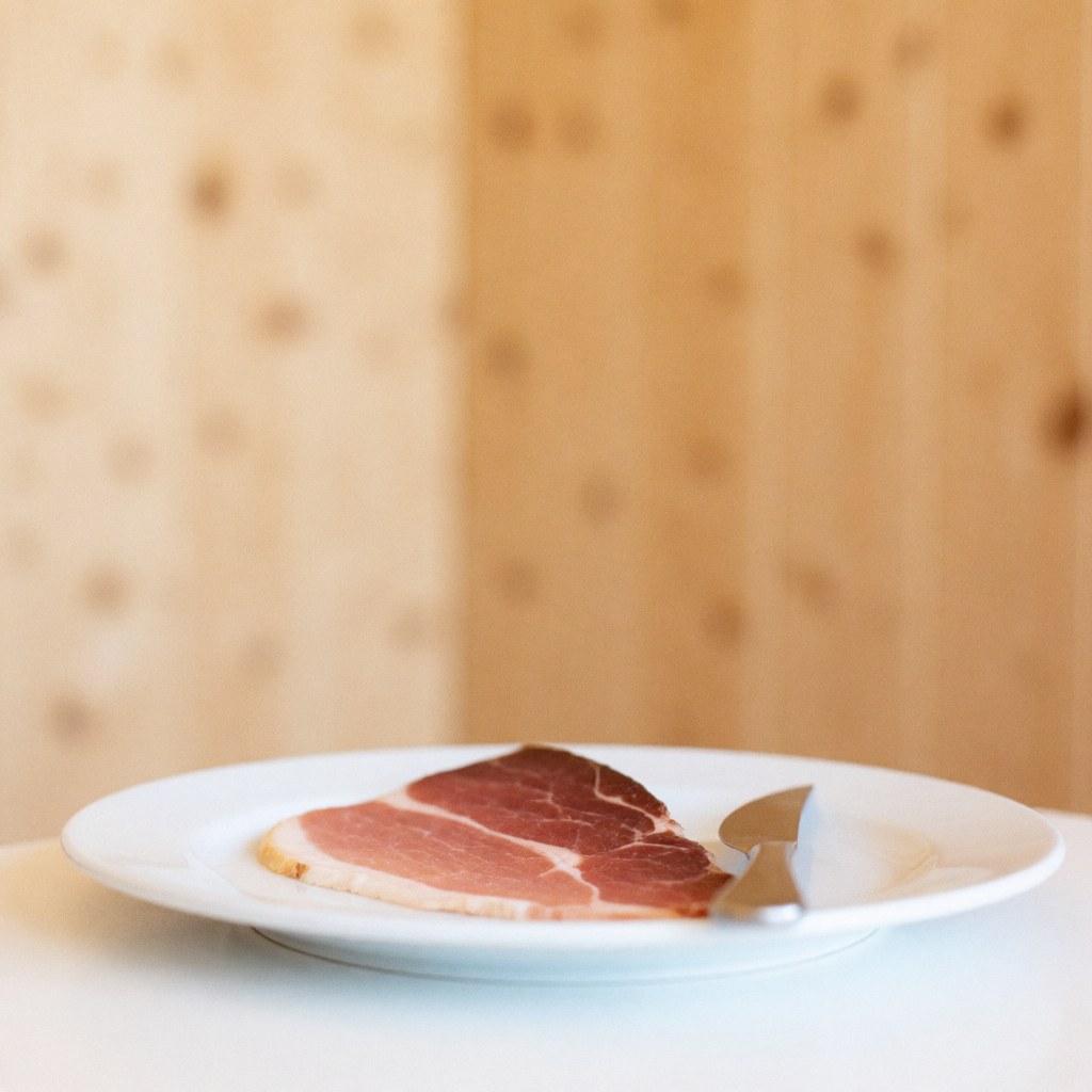 Une tranche de jambon posée sur une assiette blanche avec un couteau, sur une surface en bois à la lumière naturelle.