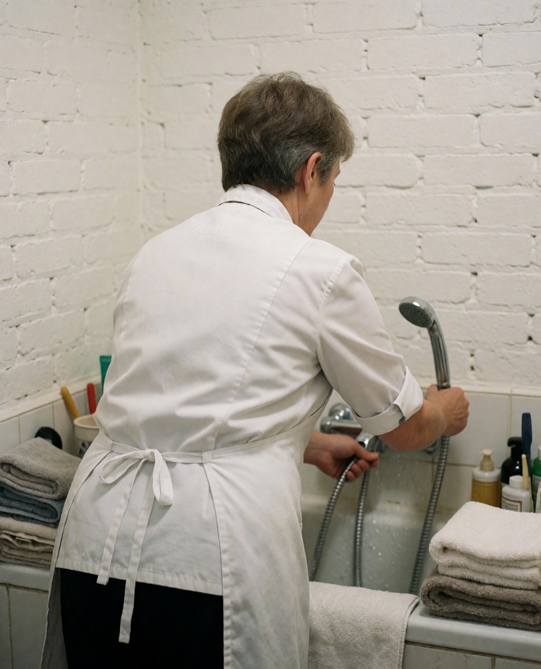 Un groomer professionnel règle la douche au-dessus d'une baignoire, avec des serviettes rangées et des murs en briques blanches, montrant un environnement propre pour le soin canin.