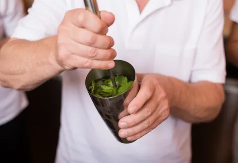 Un bartender professionnel écrase des feuilles de menthe fraîche dans un shaker métallique sur un fond flou avec des murs en terre cuite.