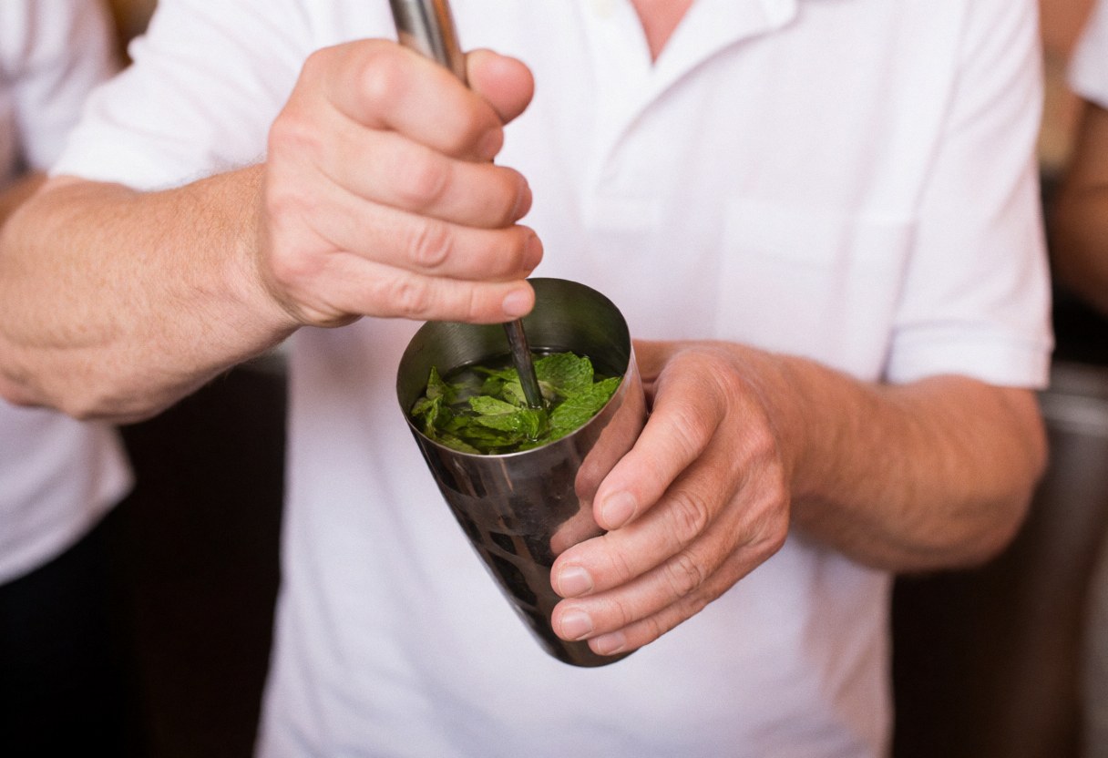 Un bartender professionnel écrase des feuilles de menthe fraîche dans un shaker métallique sur un fond flou avec des murs en terre cuite.