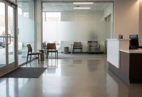 Vue d'un bureau propre et moderne avec une réception, des chaises, une table, un sol poli en béton et des parois vitrées baignées de lumière naturelle.