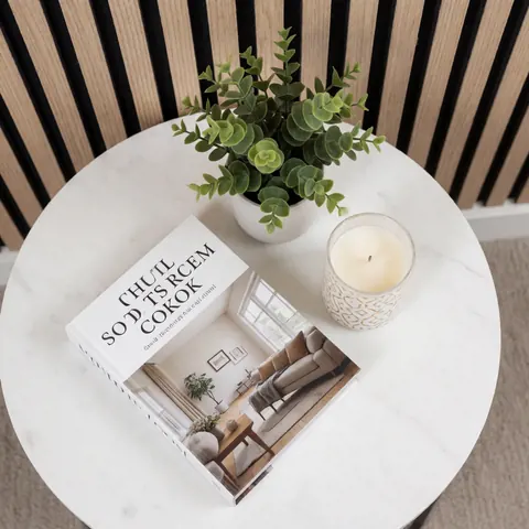 Table basse ronde en marbre blanc ornée d'un livre décoratif, d'une plante artificielle et d'une bougie, devant un mur à lamelles bois.