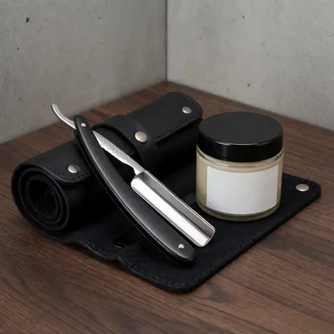 Rasoir à lame droite posé sur un étui en cuir noir, accompagné d'un pot à savon à raser, sur une surface en bois sombre et un mur de béton.