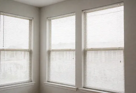Intérieur lumineux et propre avec trois grandes fenêtres aux stores blancs ouverts, murs gris clair et éclairage naturel doux.