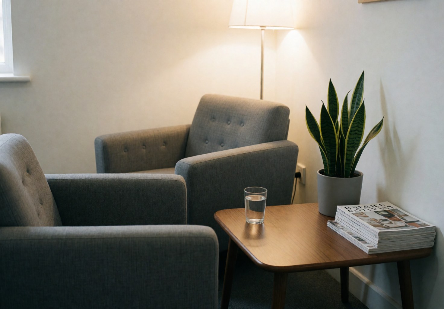 Deux fauteuils gris dans une salle d'attente de spa, avec une table en bois, une plante, un verre d'eau et des magazines. Lumière chaude et ambiance relaxante.