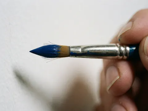 Gros plan d'un pinceau bleu tenu par une main, montrant les poils et la peinture humide sur fond blanc éclairé.