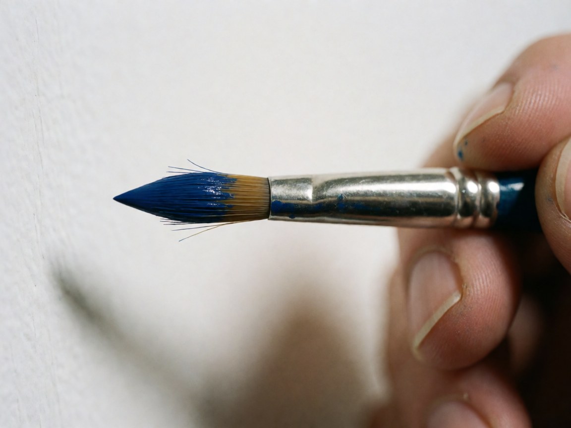Gros plan d'un pinceau bleu tenu par une main, montrant les poils et la peinture humide sur fond blanc éclairé.
