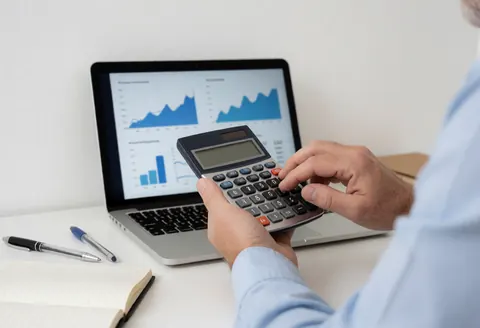 Un consultant examine des données financières sur une calculatrice tout en travaillant sur un ordinateur portable avec des graphiques, sur un bureau avec un carnet et des stylos.