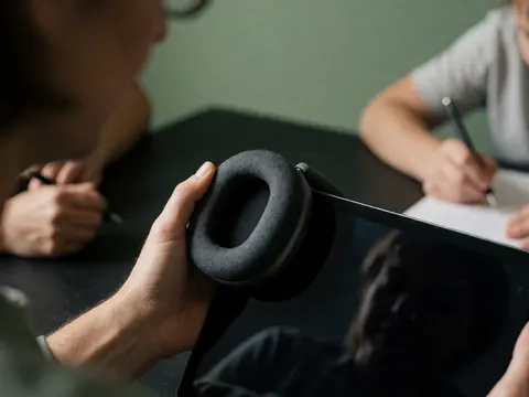 Vue rapprochée d'un casque audio et d'une tablette posés sur une surface, entourés de mains tenant des outils pédagogiques, sur un fond vert doux évoquant un environnement professionnel.