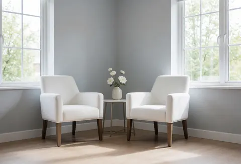 Salle de soins vide avec deux chaises blanches, une table fleurie, grandes fenêtres, murs gris et sol en bois clair.